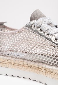 Gros plan sur une chaussure de sport avec une tige en mesh argent métallique, des lacets argentés, une languette en daim beige, une semelle intermédiaire en jute tissée et une semelle extérieure blanche rainurée.