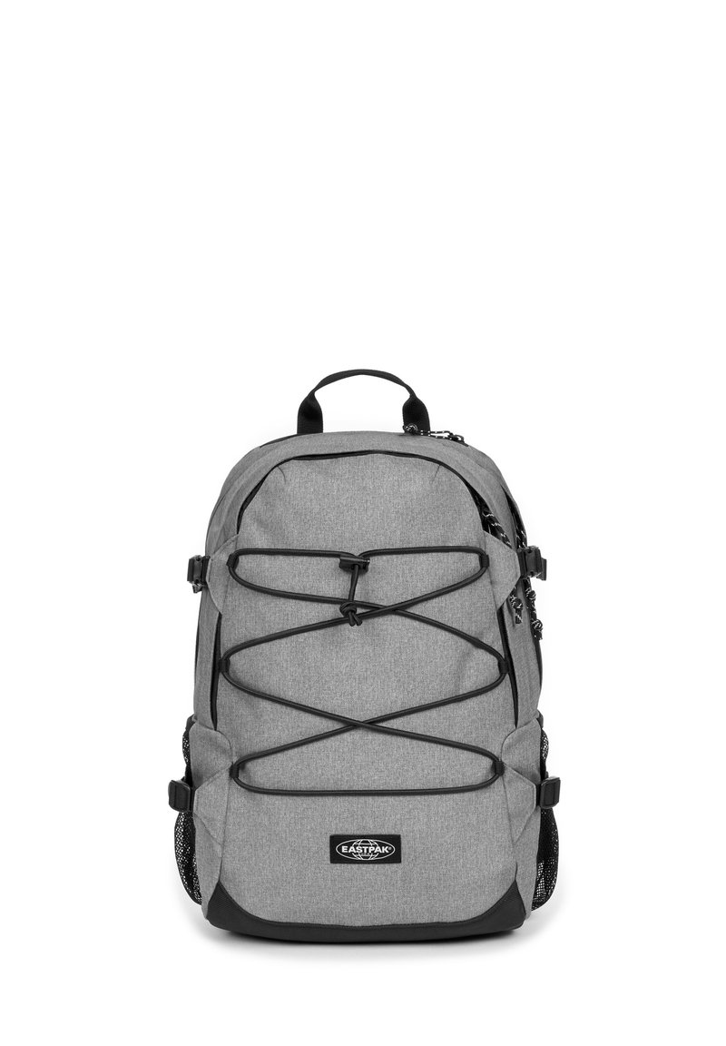 Mochila gris con un acabado texturizado, que presenta acentos negros, cuerdas elásticas en la parte frontal, doble bolsillo de malla lateral y un asa de agarre en la parte superior.