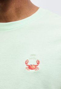 Μεντ πράσινο πουκάμισο με μικρό κεντημένο κόκκινο καβούρι κάτω από το κείμενο "OH CRAB" στο στήθος, φορεμένο από άτομο με καστανό δέρμα ορατό στον λαιμό.