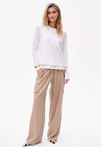 Weiße Langarmbluse und beigefarbene weit geschnittene Cargo-Hoseng mit Taschen, verziert mit einem gemusterten Kordelzug und spitzen Schuhen.