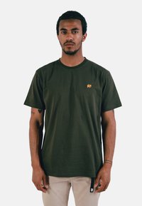 T-shirt en coton vert olive avec un petit logo d'éléphant jaune sur la poitrine, col rond et manches courtes. Texture lisse.