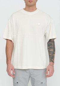 T-shirt crema a maniche corte con scollatura rotonda, caratterizzata da un logo circolare dorato sul lato sinistro del petto e un design dalla vestibilità rilassata.