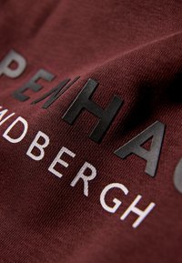 Tessuto bordeaux con finitura texture, con lettere nere in rilievo "COPENHAGEN" e bianche "AARHUS" su un capo a maniche corte.