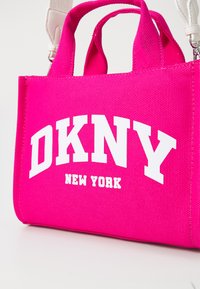 Roza platnena torba z teksturirano površino, ki vsebuje belo besedilo "DKNY NEW YORK". Ima kratke ročaje in snemljiv trak za večnamensko uporabo.