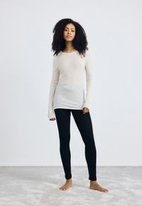 Beige lange mouwen top met een aansluitende pasvorm en een textuurstof, gecombineerd met zwarte legging. Model staat blootsvoets op een lichtgrijs tapijt.