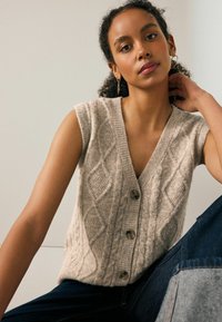 Beesid kootud без рукавов kampsun V-kaelusega, millel on teemantmustrid ja kolm esimest nööpi. Seljas tume teksapüksid.