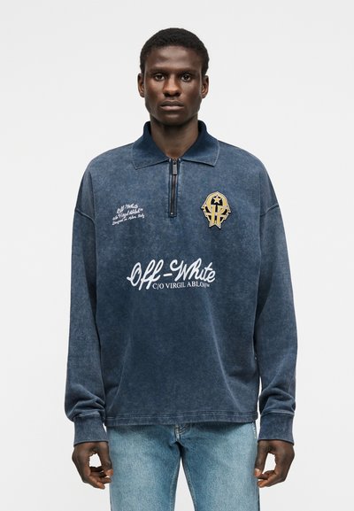 Άνδρας που φοράει ξεθωριασμένο μπλε σκούρο φούτερ με φερμουάρ στο ένα τρίτο, με το λογότυπο και το έμβλημα της Off-White, συνδυασμένο με ανοιχτόχρωμο μπλε τζιν, στέκεται μπροστά σε απλό φόντο.