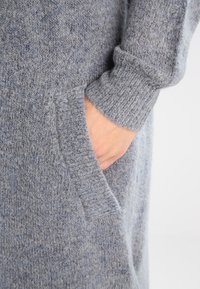 Grå stickad plagg med en mjuk textur, med en framficka och upplagda ärmar. Tyget har ett subtilt mönster av varierande nyanser.