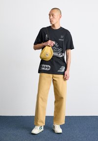 Svart t-shirt med vitt tryck, ljusbeige byxor med vida ben, gul axelremsväska och vita sneakers med gröna detaljer.