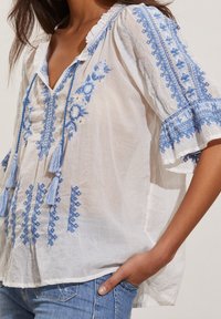 Vit broderad blus med blå blommönster och geometriska mönster, med korta, volangärmar och fransdetaljer.