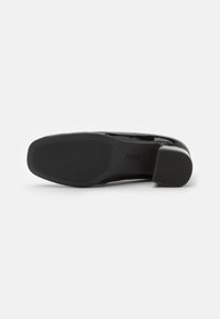 Chaussure en cuir verni noir avec un bout arrondi et un petit talon. La semelle plate présente une surface texturée pour une meilleure adhérence, avec des détails de marque.
