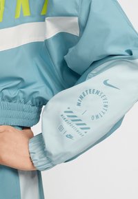 Jacheta ușor albastră are manșete elastice și talie strânsă. Partea din față are o dungă albă și Inscripții mari verzi. Mâneca prezintă detalii de branding.