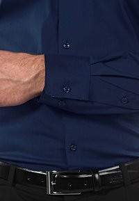 Camicia abbottonata blu navy con maniche lunghe, caratterizzata da bottoni scuri e una trama liscia, infilata in pantaloni neri con una cintura lucida.