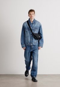Casaco de ganga com riscas brancas verticais, combinado com jeans azuis largos e sapatos pretos de slip-on. Pequena mala preta de ombro usada em banda cruzada.