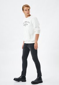 Suéter blanco con cuello redondo, detalle de texto negro, combinado con jeans ajustados oscuros y botas negras de tobillo con paneles elásticos en los laterales.