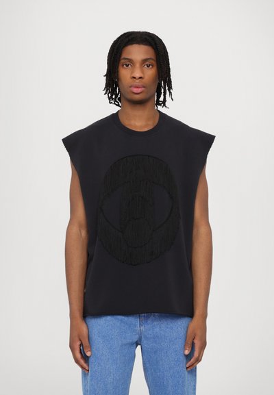 Melnā bez piedurknēm blūze ar teksturētu frizūras logo dizainu, izgatavota no mīksta auduma ar apaļu kakla izgriezumu, kopā ar gaiši ziliem džinsiem.