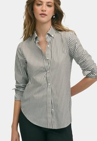 Bílé tričko s úzkými černými vertikálními pruhy, límcem na knoflíky a ohrnutými rukávy. Látka vypadá hladce a má přiléhavý střih.