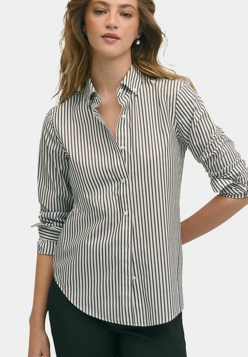 Bílé tričko s úzkými černými vertikálními pruhy, límcem na knoflíky a ohrnutými rukávy. Látka vypadá hladce a má přiléhavý střih.