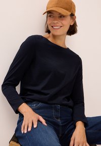 Leende kvinna med en beige keps, svart långärmad tröja och blå jeans, sittande med ena handen vilande på sitt lår mot en ljus bakgrund.