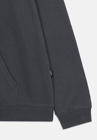 Mânca de hanorac gri închis cu un buzunar lateral, manșete ribbate și un mic eticheta de accent turcoaz aproape de margine. Textură netedă.