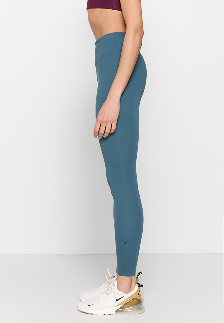 Leggings atléticos en color azul verdoso, fabricados con una tela elástica, que presentan una cinta de cintura alta y un pequeño logo en la pierna izquierda. Combinados con zapatillas blancas y doradas.