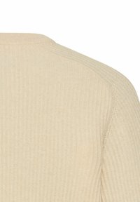 Zbliżenie na beżową tkaninę ribbed knit, ukazujące szew na ramieniu oraz teksturowane pionowe linie.