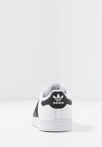 Bílé tenisky s černými detaily. Mají hladký kožený svršek, síťovinové boční panely a texturovanou gumovou podrážku. Logo Adidas na patě.