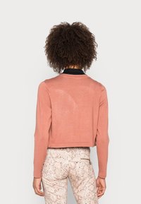 Kvinna med lockigt hår, klädd i en kort rosa tröja och ljusa byxor med ormprint, stående vänd bort mot en enfärgad bakgrund.