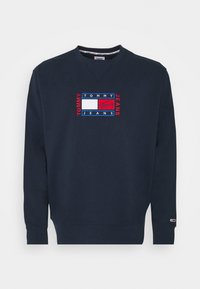 Granatowa bluza wykonana z mieszanki bawełny, z haftowanym czerwono-białym logo w formie kwadratu na piersi, ściągaczami na mankietach i dole.