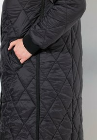 Quiltad svart ytterkläder med snedställd sömnad, utrustad med en dragkedjeficka och ribbstickade muddar, tillverkad av glänsande, lätt material.