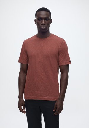 Homme aux cheveux courts portant un t-shirt à manches courtes couleur rouille avec un col rond et un pantalon noir, debout devant un fond blanc uni.