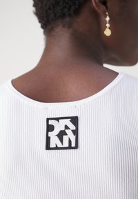 Personne à la peau foncée portant un haut blanc côtelé avec un écusson du logo DKNY noir et blanc sur le haut du dos et une boucle d'oreille pendante en perle dorée.