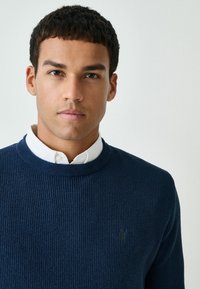 Marineblaue, gerippte Strickjacke mit Rundhalsausschnitt, die über einem weißen Hemd mit Kragen getragen wird, und mit einem kleinen gestickten Logo auf der Brust.