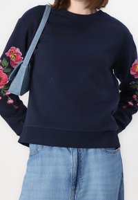 Sudadera de marina con bordado floral en rosa y verde en las mangas, puños acanalados y dobladillo. Acompañada de un pequeño bolso de mezclilla.