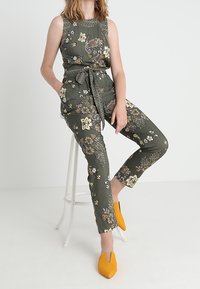 Mono floral verde con cintura ajustada y cinturón, que presenta patrones bordados y piernas ceñidas. Combinado con zapatos amarillos brillantes de punta.