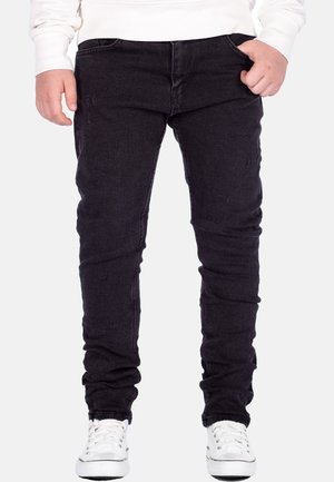 Kind trägt schwarze Slim-Fit-Jeans, weiße Sneakers und ein weißes Langarmshirt und steht mit den Händen nahe den Taschen.