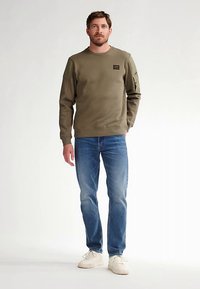 Felpa verde oliva con polsini e orlo a costine, con etichetta. Abbinata a jeans blu e sneakers bianche, in piedi su uno sfondo chiaro.