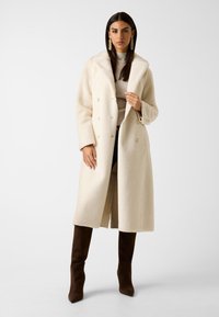 Double-breasted jas in zacht, crèmewit stof met een rijke textuur, grote kraag en goudkleurige knopen; gecombineerd met bruine kniehoge laarzen.