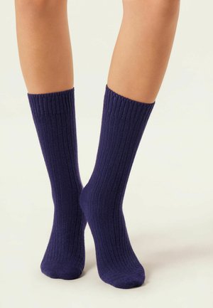 Chaussettes en laine violette avec une texture côtelée, atteignant la longueur mi-mollet. Le tissu semble doux et extensible, conçu pour le confort et la chaleur.