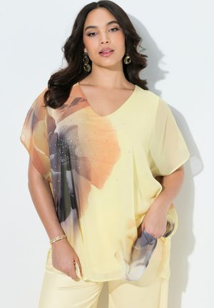 Kobieta ubrana w żółtą, luźną bluzkę z dużym kwiatowym nadrukiem i kryształkami, w połączeniu z żółtymi spodniami, pozująca na białym tle.