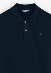 Polo bleu marine avec deux boutons blancs et petit logo tête de mort avec os croisés sur le côté gauche de la poitrine.