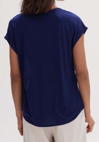 Kvinna i en marinblå kortärmad blus och ljusa byxor, sedd bakifrån mot en neutral bakgrund.