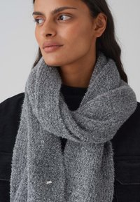 Gray knitted scarf with a soft, fuzzy texture, featuring subtle metallic threads and a small brand tag, wrapped around a dark sweater.