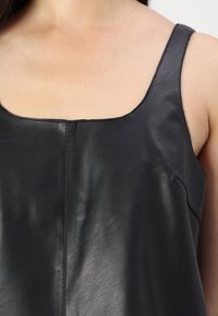 Kvinna som bär en ärmlös svart topp i konstläder med fyrkantig halsringning och synlig söm mitt fram.