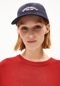 Casquette de baseball bleue marine avec le texte brodé en blanc "ARMEDANGELS SINCE 2007." Pull en tricot rouge avec un col rond et un tissu texturé.