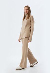 Maglione a collo alto beige e pantaloni a gamba larga abbinati in un tessuto morbido e texturizzato. Modella che indossa sneakers bianche con accenti beige.