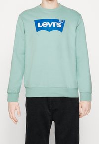 Person i en mintgrön sweatshirt med en blå Levi's-logga och svarta kordbyxor, stående mot en enfärgad bakgrund.