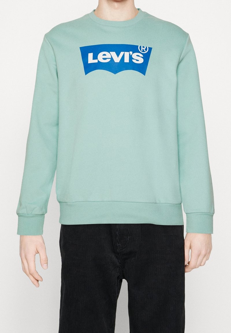 Person i en mintgrön sweatshirt med en blå Levi's-logga och svarta kordbyxor, stående mot en enfärgad bakgrund.