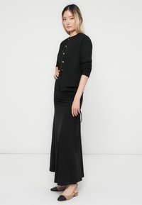 Woman standing against white background, wearing black buttoned cardigan, long black skirt, and beige flat shoes with black toes.