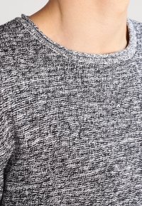 Šedý a bílý pletený svetr se strukturovaným vzorem, kulatým výstřihem a kontrastním žebrovaným lemem kolem límce.